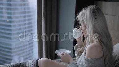 美丽的金发女人拿着陶瓷杯的茶或咖啡享受。 在床上<strong>喝茶</strong>。 看着窗户<strong>喝茶</strong>